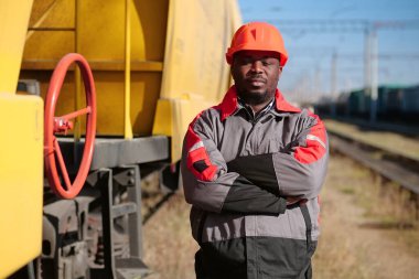 Afrikalı Amerikan demiryolu işçisi yük treni terminalinde duruyor ve kameraya bakıyor. Demiryolu işçisi üniformalı ve kırmızı şapkalı demiryolu raylarında duruyor.