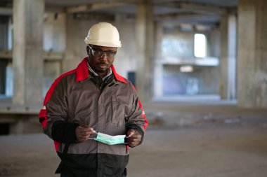 Afrikalı Amerikalı müteahhit inşaat alanında duruyor, kameraya bakıyor ve elinde tıbbi koruyucu maske tutuyor.