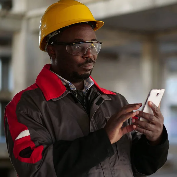 stock image African american workman in yellow hard hat with smartphone stands at construction site. Worker holds in hands smartphone