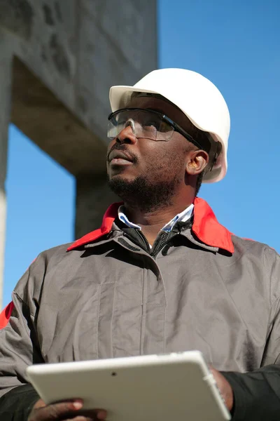 African american worker in hard hat and protective goggles with tablet computer stands at construction site and looks around