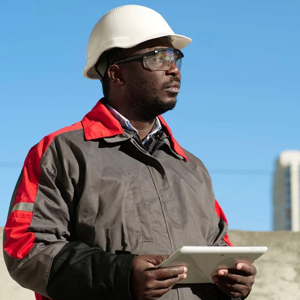 African american worker in hard hat and protective goggles with tablet computer stands at construction site