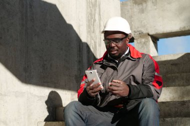 Siyah şapkalı ve koruyucu gözlüklü Afro-Amerikan işçiler inşaat alanında oturuyorlar. İşçi el ele tutuşuyor, akıllı telefon, video bağlantısı ile konuşuyor ve gülümsüyor.