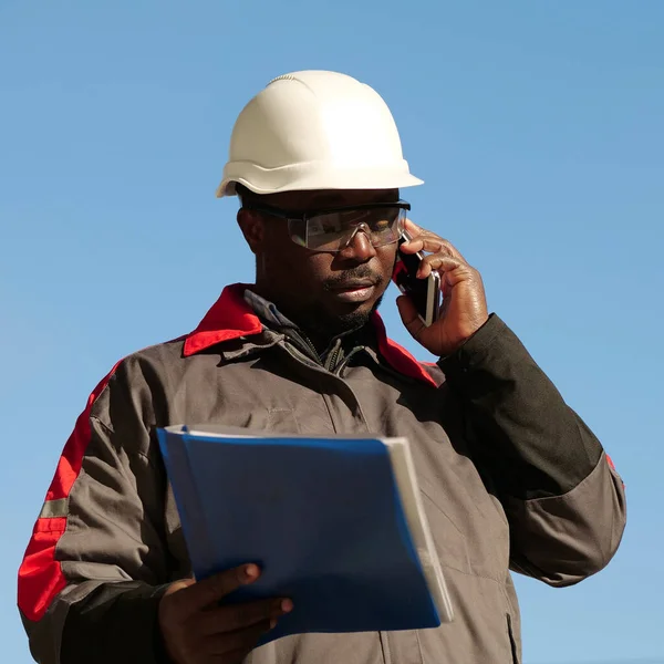 African american builder with work papers talks on smartphone. Busy african american works manager in hard hat with work documents stands at construction site and speaks on smartphone