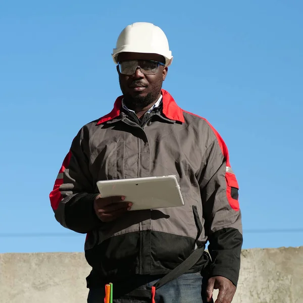 African american worker in white hard hat and protective goggles stands at construction site, holds in hands tablet computer and looks at the camera