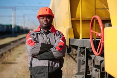 Afrikalı Amerikan demiryolu işçisi, yük treni terminalinde gözleri kapalı duruyor. Demiryolu işçisi üniformalı ve kırmızı şapkalı, kameraya bak.