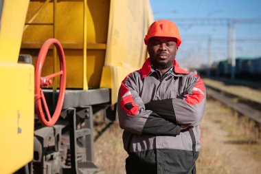 African american railway worker stands at freight train terminal. Railroad man in uniform and red hard hat look at camera