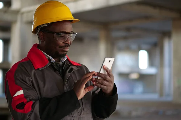 stock image African american workman in yellow hard hat with smartphone stands at construction site. Worker holds in hands smartphone