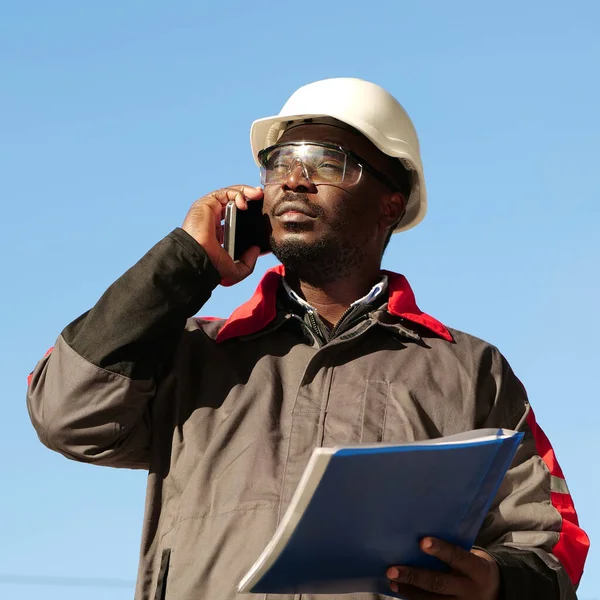 African american builder with work papers talks on smartphone. Busy african american works manager in hard hat with work documents stands at construction site and speaks on cell phone