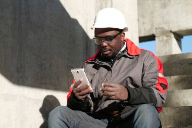 Siyah şapkalı ve koruyucu gözlüklü Afro-Amerikan işçiler inşaat alanında oturuyorlar. İşçi elinde beyaz akıllı telefon ve video bağlantısı ile konuşuyor