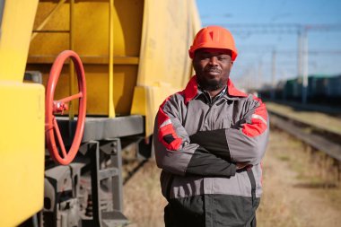 Afrikalı Amerikan demiryolu işçisi yük treni terminalinde duruyor ve kameraya bakıyor. Demiryolu işçisi üniformalı ve kırmızı şapkalı demiryolu raylarında duruyor.