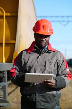 Yük treni terminalinde tablet bilgisayarı olan Afrika kökenli Amerikalı bir demiryolu işçisi. Üniformalı demiryolu işçisi ve bilgisayarlı kırmızı şapka. Demiryolu çalışanı el bilgisayarını tutuyor.