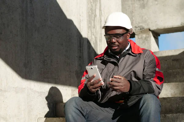 Afroamerikansk Arbetare Hård Hatt Och Skyddsglasögon Med Smartphone Sitter Byggarbetsplatsen — Stockfoto