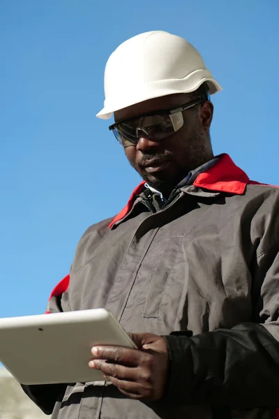 African american worker in hard hat and protective goggles with tablet computer stands at construction site