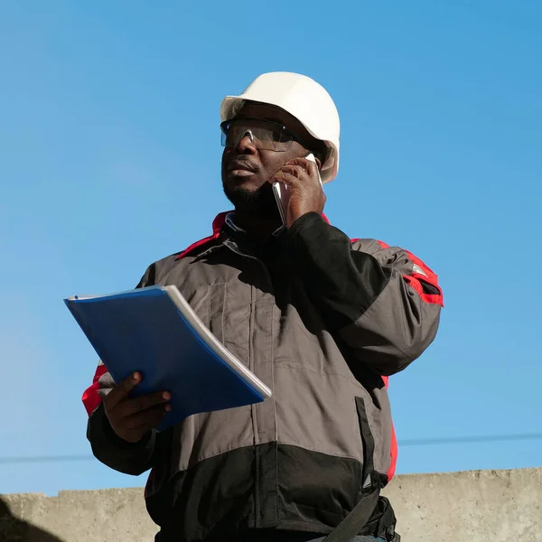 African american builder with work papers talks on smartphone. Busy african american works manager in hard hat with work documents stands at construction site and speaks on smartphone