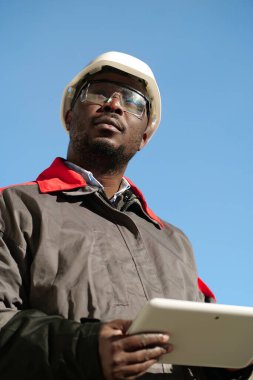 Siyah şapkalı, koruyucu gözlüklü, tablet bilgisayarlı Afro-Amerikan işçiler inşaat sahasında durup etrafa bakıyorlar.