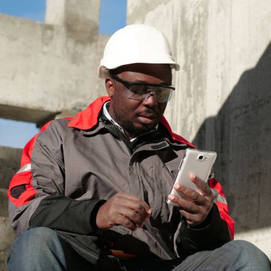 Siyah şapkalı ve koruyucu gözlüklü Afro-Amerikan işçiler inşaat alanında oturuyorlar. İşçi akıllı telefonuna bakıyor.