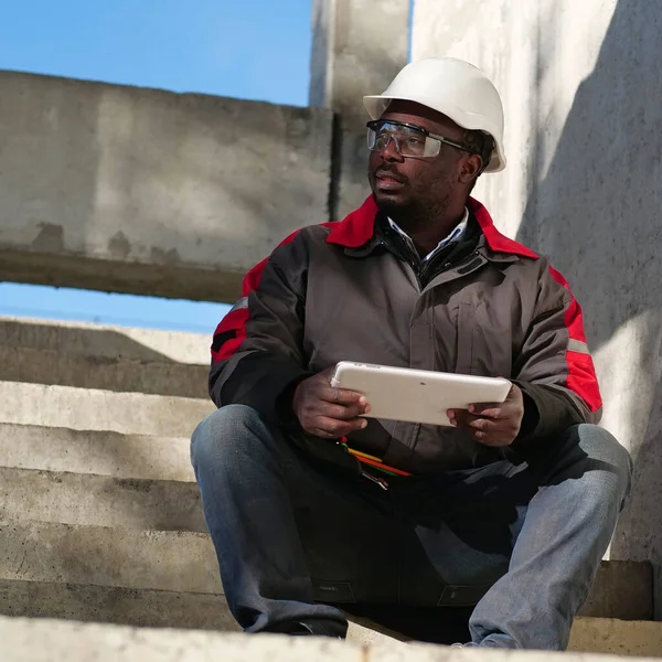 Beyaz başlıklı yarı kapalı gözlerle yorgun Afro-Amerikan işçisi şantiyede oturuyor ve tableti elinde tutuyor.