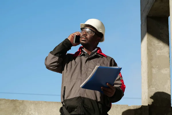 African american builder with work papers talks on smartphone. Busy african american works manager in hard hat with work documents stands at construction site, speaks on smartphone and looks at the sun