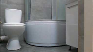 Tuvalet odası içi kapalı kapaklı beyaz klozet, duş kabini, lavabo ve gri duvarın altındaki dolap ve yer döşemeleri.