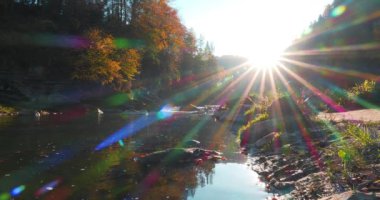 Dağlarda gün batımı, güneş ışığını yansıtan nehir, havada uçan böcekler. Sarı sonbahar ağaçları nehir kıyısında yetişir. Çok uzun renkli ışınları olan bir güneş, kapalı açıklıklı bir kamera. Sinema 4K 60fps video