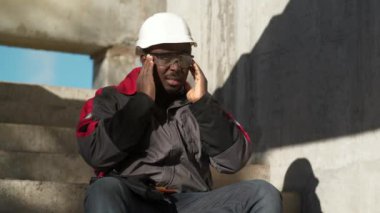 Slow Motion Afrikalı Amerikalı işçiler inşaat alanının beton merdivenlerinde oturuyor. Kendini iyi hissetmiyor ve başı ağrıyor. Gözlüklerini çıkarıp kafasına masaj yapıyor.