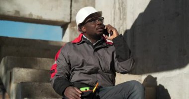 Afrikalı Amerikalı işçiler akıllı telefondan konuşur. Beyaz şapkalı ve koruyucu gözlüklü inşaatçı inşaat alanındaki beton merdivenlerde oturuyor ve cep telefonuyla konuşuyor.