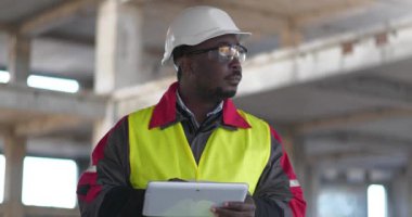 Siyah şapkalı ve koruyucu gözlüklü Afrikalı Amerikalı işçi inşaat alanında duruyor ve tablet bilgisayarına notlar yazıyor.