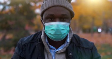Tıbbi koruyucu maskeli ve kışlık şapkalı Afro-Amerikan bir adam sonbahar parkında oturup kameraya bakar.