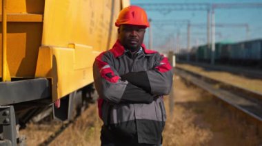 Afrikalı Amerikan demiryolu işçisi yük treni terminalinde duruyor ve kameraya bakıyor. Demiryolu işçisi üniformalı ve kırmızı şapkalı demiryolu raylarında duruyor.