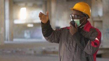 Yavaş Hareketli Afrikalı Amerikalı işçiler sarı şapkalı, gözlüklü ve tıbbi koruyucu maskeli inşaat alanında duruyor ve taşınabilir radyo vericisiyle konuşuyor ve elleriyle işaret ediyor.