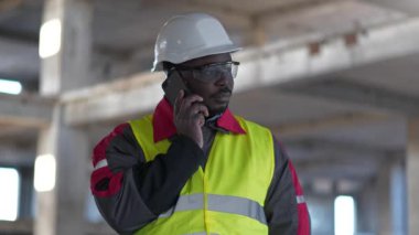Slow Motion Afrikalı Amerikalı inşaatçı akıllı telefondan konuşuyor. İnşaat alanındaki işletme müdürü cep telefonuyla konuşuyor