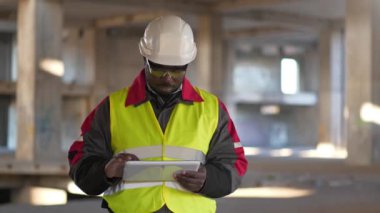 Siyah şapkalı ve koruyucu gözlüklü Afrikalı Amerikalı işçiler inşaat alanında duruyor ve tablet bilgisayarla video bağlantısıyla konuşuyor.
