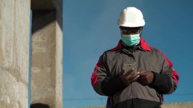 Siyah şapkalı ve koruyucu maskeli Afrikalı Amerikalı işçi inşaat alanında duruyor ve video bağlantısıyla akıllı telefondan konuşuyor.