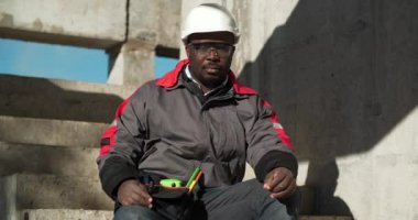 Afrikalı Amerikalı işçiler inşaat alanında merdivenlerde oturur ve kameraya bakar. Sinema 4K 60fps video