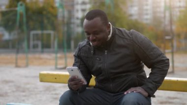 Afrikalı Amerikalı bir adam spor sahasında oturup video bağlantısıyla akıllı telefondan konuşuyor.