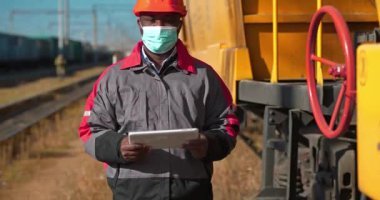 Tıbbi maske takan yük treni terminalinde çalışan Afro-Amerikan demiryolu işçisi el bilgisayarını tutuyor ve kameraya bakıyor. Yük istasyonunda tabletli demiryolu trafiği müfettişi.