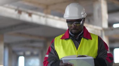 Slow Motion Afrikalı Amerikalı işçi beyaz şapka ve koruyucu gözlüklü inşaat alanında duruyor ve tablet bilgisayarına notlar yazıyor.
