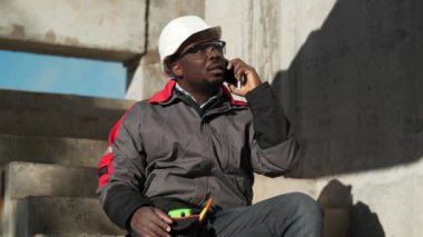 Slow Motion Afrikalı Amerikalı işçi akıllı telefondan konuşuyor. Beyaz şapkalı ve koruyucu gözlüklü inşaatçı inşaat alanındaki beton merdivenlerde oturuyor ve cep telefonuyla konuşuyor.