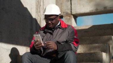 Afrikalı Amerikalı işçiler inşaat alanında merdivenlerde oturur ve video bağlantısıyla akıllı telefondan konuşur.
