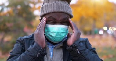 Tıbbi koruyucu maskeli ve kış şapkalı Afro-Amerikan adam kendini iyi hissetmiyor ve başı ağrıyor, kafasına masaj yapıyor.
