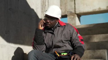 Afrikalı Amerikalı işçiler akıllı telefondan konuşur. İnşaatçı inşaat alanındaki beton merdivenlerde oturur ve cep telefonuyla konuşur, etrafa bakar.