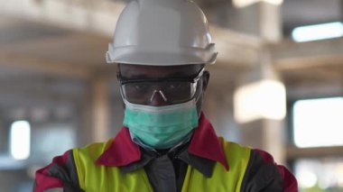 Tıbbi koruyucu maskeli Afrikalı Amerikalı müteahhit inşaat alanında duruyor ve kameraya bakıyor, yakın çekim.