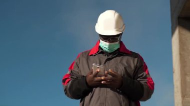 Siyah şapkalı ve tıbbi koruyucu maskeli Afrikalı Amerikalı inşaatçı inşaat sahasında duruyor ve akıllı telefondan video bağlantısıyla konuşuyor.