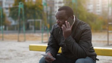 Afrika kökenli Amerikalı bir adam spor sahasında oturuyor, akıllı telefonla konuşuyor ve elinde ikinci bir akıllı telefon tutuyor.