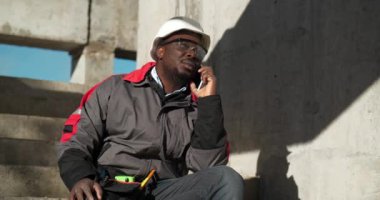 Afrikalı Amerikalı müteahhit akıllı telefondan konuşuyor. İnşaatçı inşaat alanındaki beton merdivenlerde oturur ve cep telefonuyla konuşur, etrafa bakar.