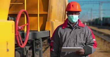 Elinde Tablet PC ile tıbbi maske takan Afro-Amerikan demiryolu işçisi yük treni terminalinde kameraya bakıyor. Yük istasyonunda tablet bilgisayarlı demiryolu trafiği müfettişi