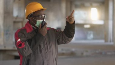 Siyah şapkalı, gözlüklü ve tıbbi koruyucu maskeli Afro-Amerikan işçiler inşaat alanında duruyor ve taşınabilir radyo vericisiyle konuşuyor.
