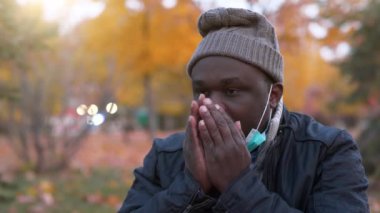 Slow Motion hasta Afro-Amerikan kışın şapkalı Sonbahar Şehri parkında tıbbi koruyucu maskesini çıkarıyor, hapşırıyor ve öksürüyor.