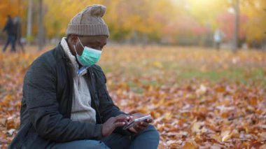Ters Zaman Yavaş Hareketi, tıbbi koruyucu maskeli ve kışlık şapkalı Afrikalı Amerikalı güneşli sonbahar parkında sarı yaprakların üzerinde oturuyor ve akıllı telefonlarla çalışıyor.