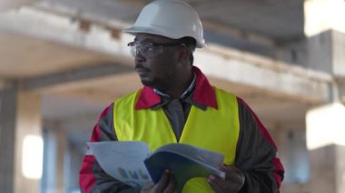 Siyah şapkalı ve koruyucu gözlüklü Afrikalı Amerikalı müteahhit inşaat sahasında duruyor.